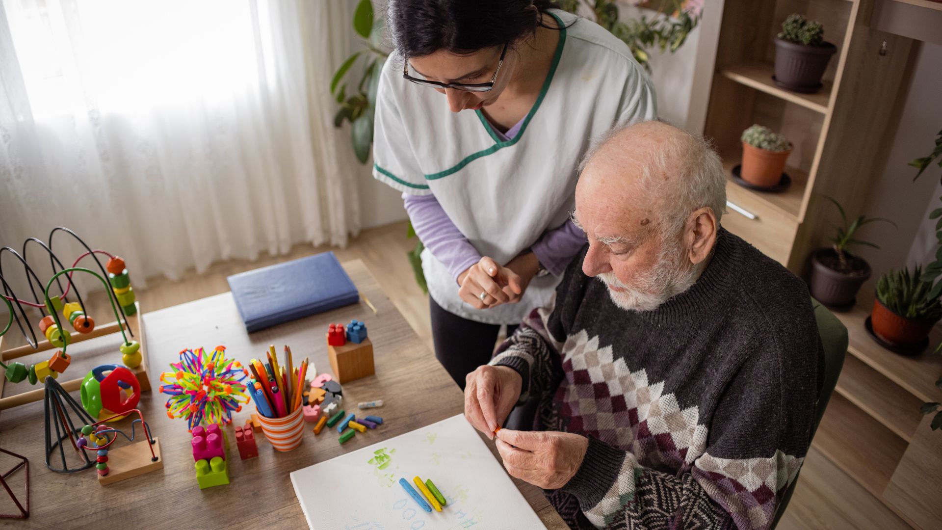 Caregiver assists elderly man with art therapy using colorful craft supplies