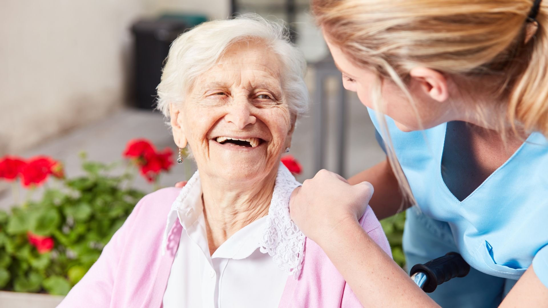 Elderly woman laughing with caregiver near red flowers